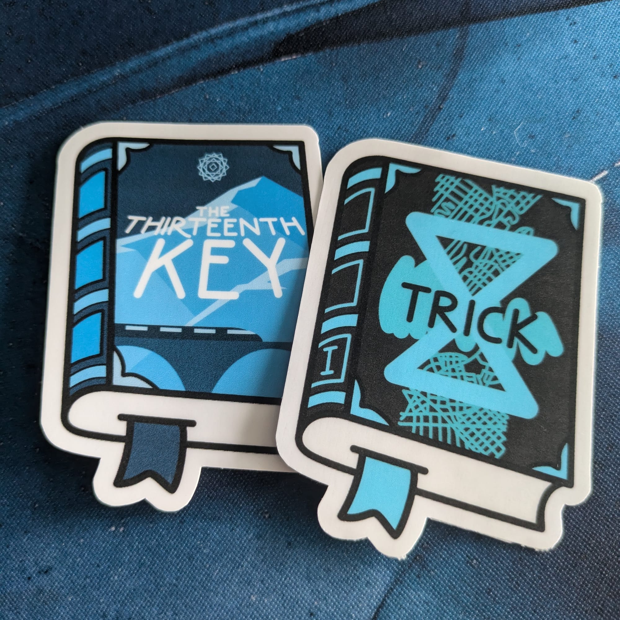 Two book-shaped stickers with white borders, matching the covers of The Thirteenth Key by Cara Nox and Trick by Cara Nox. Both are laying flat, slightly overlapping each other, on a blue marbled desk mat.